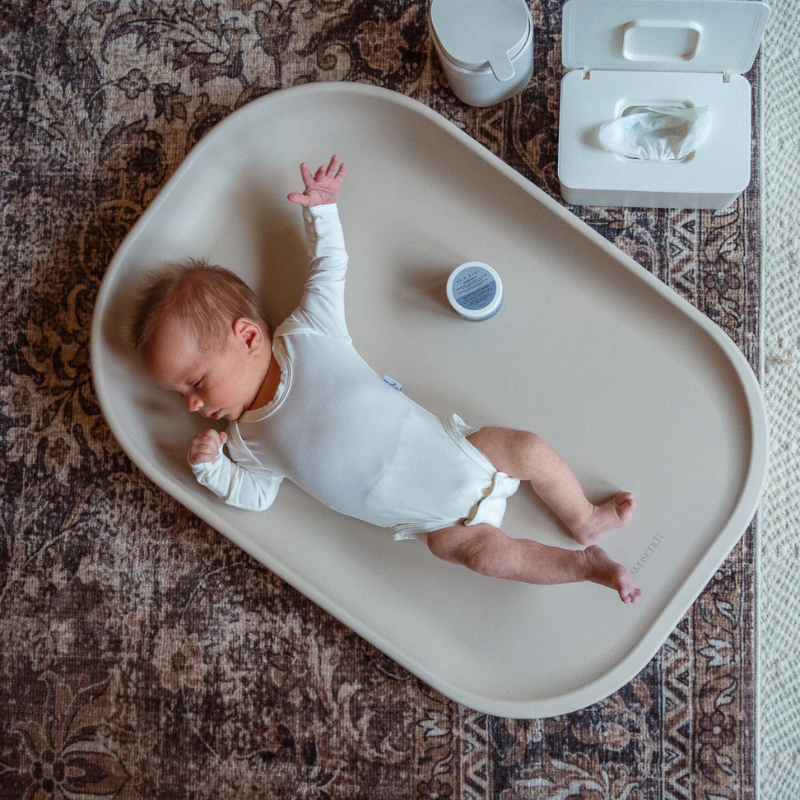 Newborn baby lying on a white changing pad with a diaper and wipes nearby on a patterned rug.