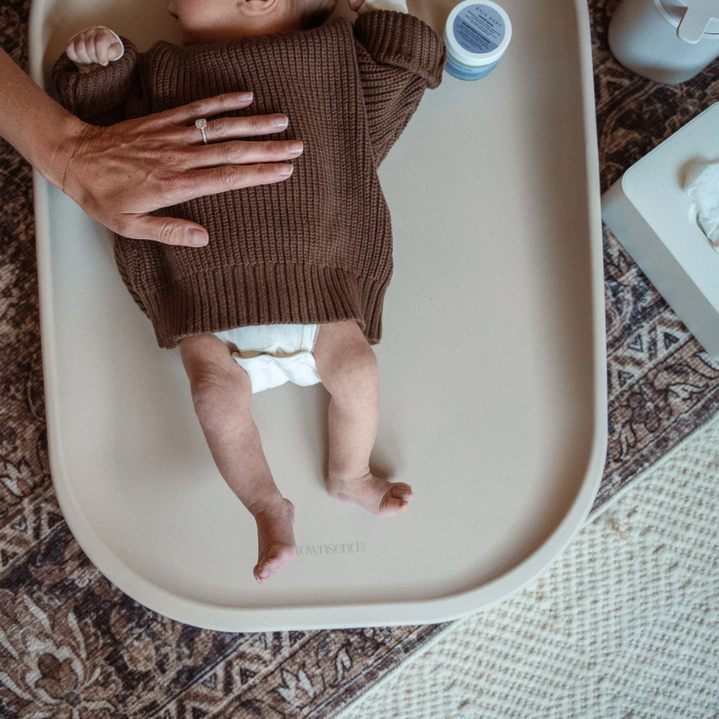 Baby in a brown outfit lying on a white tray with a textured rug underneath