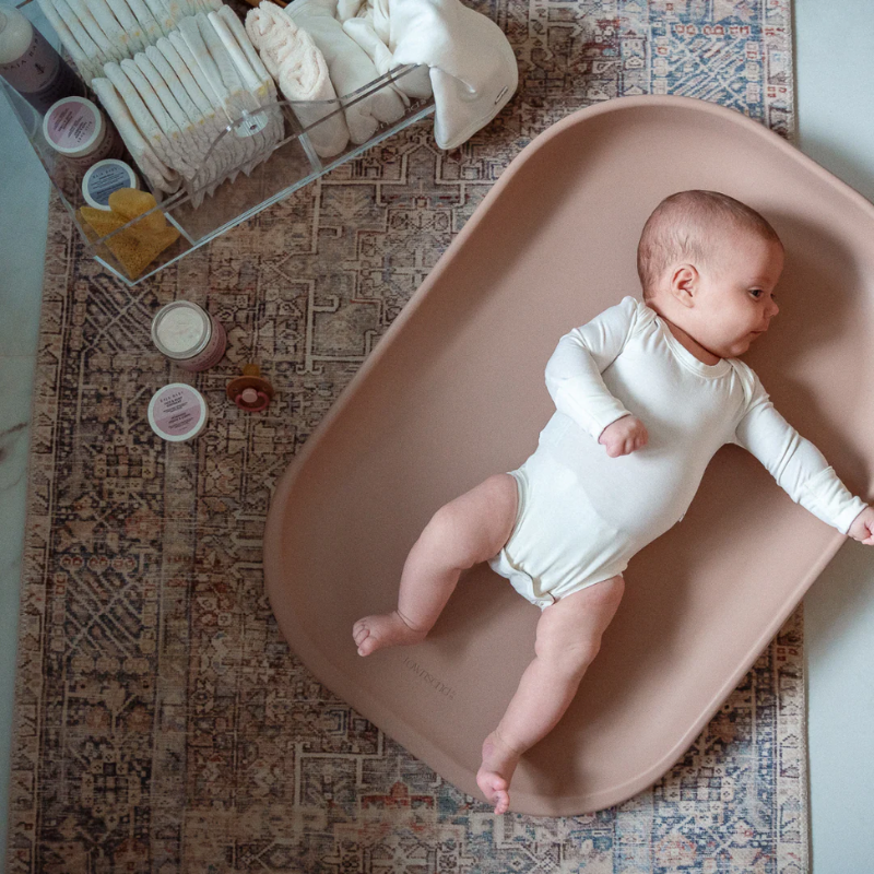 Baby in a white onesie lying on a pink waterproof contoured changing pad with baby care items around.